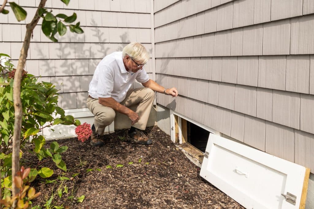 Man inspecting the base of shingle-style siding and a crawl space panel, the kind of exterior check a Louisiana insurer may request