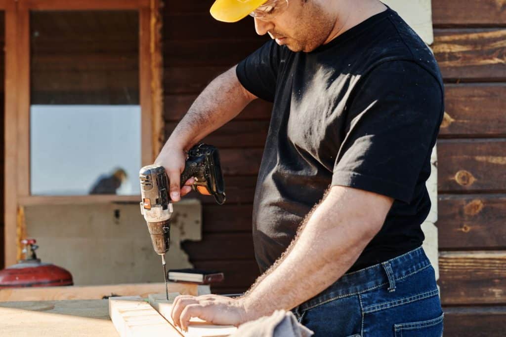Worker in a hard hat and safety glasses drilling a board next to a house with dark horizontal wood siding